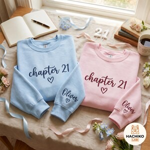 Peut inclure: Un sweat-shirt bleu clair et un sweat-shirt rose pâle, tous deux ornés du texte brodé "chapter 21" avec un cœur sur la poitrine et le prénom "Oliver" avec un cœur sur le poignet de la manche, posés sur une surface en tissu neutre.