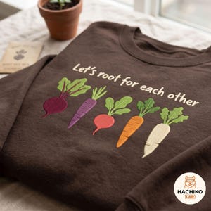 May include: A dark brown crewneck sweatshirt featuring an embroidered graphic of five colourful root vegetables including a beet, purple carrot, radish, orange carrot, and white parsnip, with the text Let's root for each other in white.