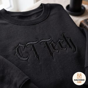 Può includere: Una felpa nera a girocollo con la scritta CT Tech ricamata in filo nero in un carattere in stile gotico. Il capo presenta un colletto a coste e maniche lunghe. Un piccolo logo circolare con un cane riporta la scritta HACHIKO LAB.