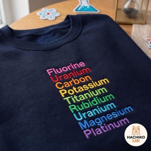 Könnte beinhalten: Ein marineblaues Sweatshirt mit gesticktem Text in einem Regenbogenverlauf. Die aufgelisteten Wörter sind Fluorine, Uranium, Carbon, Potassium, Titanium, Rubidium, Uranium, Magnesium und Platinum. Die Stickerei ist mittig auf der Brust platziert.