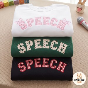 Può includere: Tre felpe girocollo impilate verticalmente in bianco, verde scuro e nero. Ognuna presenta la parola SPEECH in grandi lettere ricamate a motivi con piccoli fiocchi alle estremità. La felpa bianca ha lettere a quadretti rosa, quella verde lettere a pois color crema e quella nera lettere a quadretti rossi.