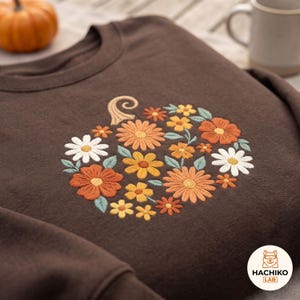 May include: A dark brown crewneck sweatshirt featuring an embroidered pumpkin shape composed of various flowers in shades of orange, yellow, and white, accented with small green leaves and a curved stem. The HACHIKO LAB logo is in the corner.