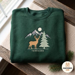 Puede incluir: Una sudadera de cuello redondo en color verde bosque oscuro con un bordado que muestra un ciervo beige junto a un pino verde claro, con montañas grises y una luna blanca de fondo. La palabra TERRASEN está bordada debajo del ciervo.