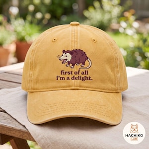 Op de afbeelding: Een mosterdgele baseballpet met een geborduurde afbeelding van een bruine opossum en de tekst "first of all i'm a delight." in donkerbruine letters. De pet heeft een gestructureerde kroon, een gebogen klep en een knoopje aan de bovenzijde.