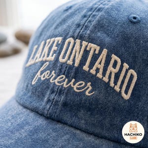 May include: A blue denim baseball cap featuring the embroidered text LAKE ONTARIO in a bold, capitalised sans-serif font, with the word forever written below in a smaller, cursive script. The embroidery is in a light cream colour.