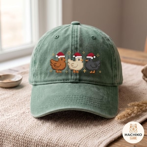 May include: A sage green baseball cap featuring a pixel art graphic of three ducks wearing red and white Santa hats. The ducks are brown, cream, and dark grey, arranged in a row with small yellow star accents above and below them.