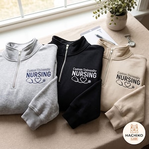May include: Three quarter-zip sweatshirts in light gray, black, and beige. Each features embroidered text reading Custom University NURSING above a stethoscope shaped into a heart. The garments have silver-toned zippers and ribbed cuffs.