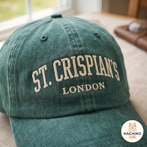 Op de afbeelding: Een donkergroene baseballpet met een gewassen, textuurrijke stoffen afwerking. De voorkant is voorzien van geborduurde crèmekleurige tekst die ST. CRISPIAN'S LONDON spelt in een vetgedrukt, gebogen schreeflettertype met een knoop.