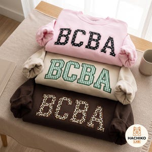 May include: Three long-sleeved crew-neck sweatshirts stacked vertically. The top is light pink with black and white polka dot letters spelling BCBA. The middle is beige with green and white gingham letters spelling BCBA. The bottom is dark brown with leopard print letters spelling BCBA.