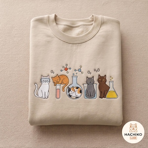 May include: A beige crewneck sweatshirt featuring a graphic of five cats interacting with laboratory glassware and chemical formulas. The design includes a white cat with goggles, a sleeping orange cat, a calico cat, a grey cat, and a brown cat.