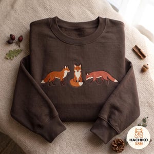 Könnte beinhalten: Ein dunkelbraunes Sweatshirt mit Rundhalsausschnitt und einer Stickerei von drei Füchsen in verschiedenen Posen in der Mitte. Die Füchse sind in Orange-, Weiß- und Schwarztönen dargestellt. Das Sweatshirt hat lange Ärmel und einen gerippten Ausschnitt.