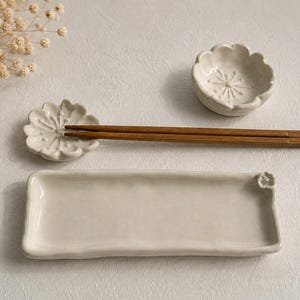 Puede incluir: Un juego de vajilla de cerámica color hueso que incluye una bandeja rectangular con un pequeño detalle floral, dos soportes con forma de flor y un par de palillos de madera marrón. Las piezas presentan un acabado liso, brillante y bordes.