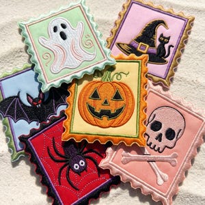 May include: A collection of six square embroidered patches with scalloped edges, featuring Halloween designs including a white ghost, a black cat with a witch hat, a bat, a jack-o'-lantern, a purple spider on a web, and a skull with crossbones.