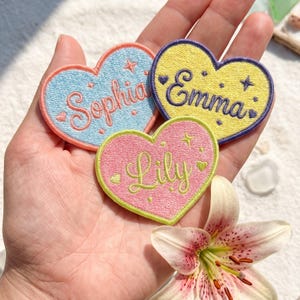 May include: Three heart-shaped embroidered patches featuring names in cursive script. One light blue patch reads Sophia, one yellow patch with a purple border reads Emma, and one pink patch with a lime green border reads Lily. Each includes small star and heart accents.