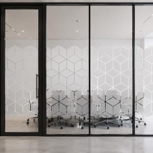 Può includere: Un set di pareti divisorie per ufficio in vetro con un motivo geometrico a cubi bianco satinato. I pannelli sono incorniciati in metallo nero e includono una maniglia nera abbinata, creando una barriera semitrasparente per una sala.
