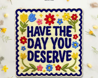 Puede incluir: Un parche bordado cuadrado con fondo color crema y borde festoneado azul oscuro. El texto "HAVE THE DAY YOU DESERVE" está bordado en letras azul oscuro, rodeado de coloridas flores bordadas en tonos rojo, amarillo y azul.