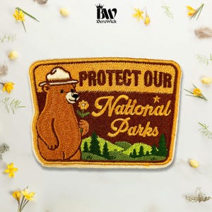 May include: A rectangular embroidered patch featuring a brown bear wearing a park ranger hat and holding a yellow flower. The text reads "PROTECT OUR National Parks" in brown and yellow thread, set against a landscape of green pine trees.