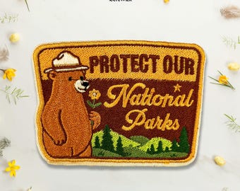 Puede incluir: Un parche bordado rectangular que muestra un oso pardo con un sombrero de guardabosques y una flor amarilla. El texto "PROTECT OUR National Parks" está bordado en hilo marrón y amarillo sobre un paisaje de pinos de color verde.