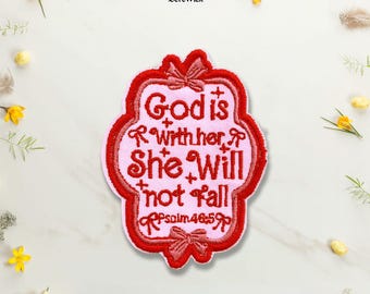 Puede incluir: Un parche bordado rosa con un borde festoneado rojo y lazos rojos en la parte superior e inferior. El texto dice God is with her, She will not fall, Psalm 46:5 en hilo rojo, acentuado con pequeñas cruces rojas sobre un fondo de tela rosa claro.