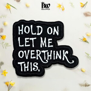 May include: A black embroidered patch with a white stitched border and the white text "HOLD ON LET ME OVERTHINK THIS." The patch has an irregular, organic shape and features a clean, bold font style that stands out against the dark fabric background.