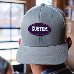 May include: A grey baseball cap with a white mesh back and a flat brim. The front features an oval-shaped navy blue patch with a red border and the word CUSTOM printed in bold white capital letters across the centre of the patch.