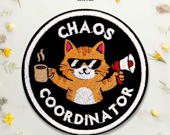 Puede incluir: Un parche bordado circular con fondo negro y borde blanco. Presenta un gato atigrado naranja con gafas de sol negras, sosteniendo una taza de café humeante en una pata y un megáfono rojo y blanco en la otra, con el texto CHAOS COORDINATOR en color blanco.