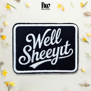 May include: A rectangular black embroidered patch with a white border. The patch features the white embroidered text "Well SHEEYIT" in a stylised cursive script. The design is centred on a plain background with small yellow floral accents.