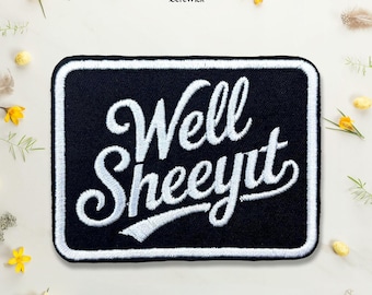Puede incluir: Un parche rectangular bordado en negro con un borde blanco. El parche presenta el texto bordado en blanco "Well SHEEYIT" en una escritura cursiva estilizada. El diseño está centrado sobre un fondo liso con pequeños detalles florales amarillos.