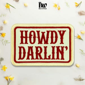May include: A rectangular embroidered patch featuring a cream-coloured background with a thick red border and a gold-coloured outer edge. The text HOWDY DARLIN' is stitched in a bold, red, serif font across the centre of the patch.