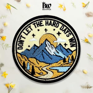 May include: A circular embroidered patch featuring a mountain landscape with a yellow sun, blue peaks, and a winding path. The text DON'T LET THE HARD DAYS WIN is stitched in black along the top border. The design includes small white clouds and stars.
