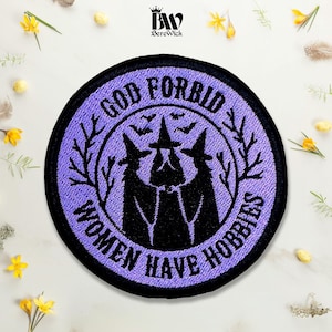 May include: A circular embroidered patch with a lavender background and a black border. The design features three black silhouettes of witches in pointed hats, surrounded by bare tree branches and bats, with the text GOD FORBID WOMEN HAVE HOBBIES.