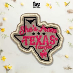 May include: A tan embroidered patch shaped like the state of Texas with a black border. It features the text 'She's from TEXAS I Can Tell' in bright pink thread, a black longhorn silhouette, three small black stars, and a pink bow graphic.