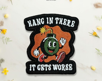 Puede incluir: Un parche bordado que presenta un personaje de bomba de dibujos animados con cara verde, guantes blancos y zapatos rojos. La bomba está sobre un fondo naranja con bordes negros. El texto dice HANG IN THERE arriba e IT GETS WORSE abajo.