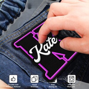 May include: A black and purple embroidered patch featuring the name Kate in white script lettering. The patch is shaped like a large letter K with a purple border and is placed on blue denim fabric. Four icons at the bottom indicate features.