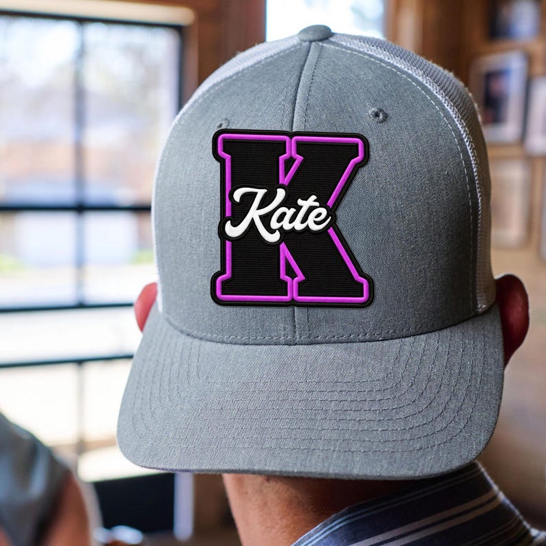 May include: A grey baseball cap with a white mesh back and a structured front panel. Centered on the front is a large black letter K with a bright pink outline. The name Kate is written in white cursive script across the centre of the letter K.