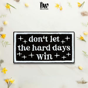 May include: A rectangular black embroidered patch with a white border and white stitched text that reads "don't let the hard days win." The design features small white four-pointed star accents scattered around the text on the black fabric.