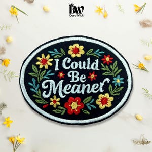 May include: An oval-shaped black embroidered patch featuring the text I Could Be Meaner in light blue script. The design is surrounded by a colourful floral border with red, yellow, and blue flowers and green leaves, finished with a light blue border.