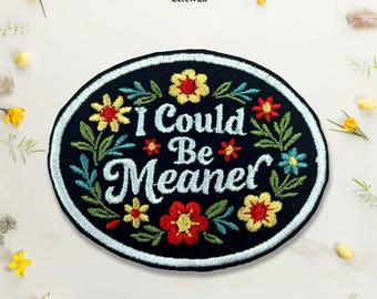 Puede incluir: Un parche bordado negro de forma ovalada con el texto I Could Be Meaner en letra cursiva azul claro. El diseño está rodeado por un borde floral colorido con flores rojas, amarillas y azules y hojas verdes, acabado con un borde azul claro.