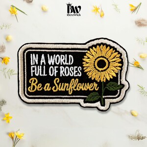 May include: A rectangular embroidered patch with a black background and a white border. It features the white text IN A WORLD FULL OF ROSES and the yellow script text Be a Sunflower, accompanied by a yellow sunflower with a green stem and leaves.