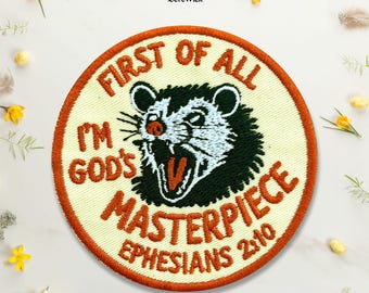 Puede incluir: Un parche bordado circular con fondo blanco hueso y borde naranja quemado. Presenta una zarigüeya en blanco y negro con la boca abierta y el texto "FIRST OF ALL I'M GOD'S MASTERPIECE EPHESIANS 2:10" en letras de color naranja quemado.