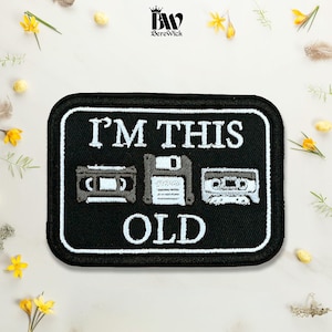 May include: A rectangular black embroidered patch with a white border. The text reads I'M THIS OLD in white capital letters. Between the words are embroidered icons of a VHS tape, a floppy disk, and a cassette tape in shades of grey and white.