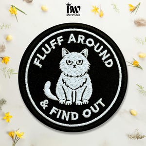 May include: A circular black embroidered patch featuring a white illustration of a grumpy cat in the center. The text "FLUFF AROUND & FIND OUT" is stitched in white capital letters around the cat, following the circular border of the patch.
