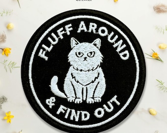 Puede incluir: Un parche bordado negro de forma circular que presenta una ilustración blanca de un gato gruñón en el centro. El texto "FLUFF AROUND & FIND OUT" está bordado en letras mayúsculas blancas alrededor del gato, siguiendo el borde circular.
