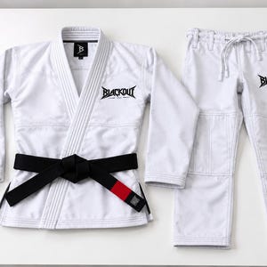 May include: A white Brazilian Jiu-Jitsu gi set consisting of a long-sleeved jacket and matching pants. The jacket features a black belt with a red tip and the text BLACKOUT embroidered on the chest. The pants also display the text BLACKOUT.