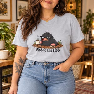 Könnte beinhalten: Ein hellgraues Kurzarm-T-Shirt mit einer Grafik eines schwarzen Maulwurfs, der eine Sonnenbrille trägt, eine dampfende weiße Kaffeetasse hält und aus einem braunen Erdhügel hervorschaut. Der Text "Woo to the Hoo" ist darunter in Schwarz gedruckt.