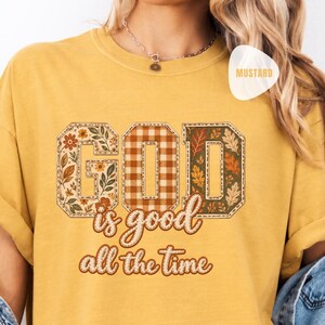 May include: A mustard yellow short-sleeve t-shirt featuring the text GOD in large block letters with floral, plaid, and leaf patterns. Below the block letters, the cursive text reads is good all the time in a brown outline and fill.