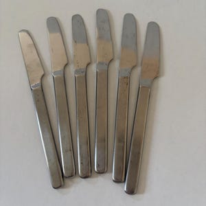 May include: A set of six silver-toned stainless steel dinner knives arranged in a fan pattern. Each knife features a smooth, flat handle and a slightly curved blade with a serrated edge, showing a consistent metallic finish across all pieces.