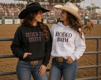 May include: A black hoodie and a white hoodie, each featuring the text "RODEO BUM" in a multicolored striped pattern across the chest. Both hoodies include a drawstring hood and a front kangaroo pocket, paired with blue denim jeans and cowboy hats.