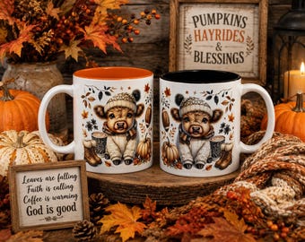 May include: Two white ceramic mugs featuring a cartoon highland cow wearing a knit hat and scarf, surrounded by autumn leaves, pumpkins, and footballs. One mug has an orange interior, and the other has a black interior. Both mugs display identical graphics.
