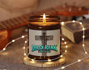 May include: A lit candle in a brown glass jar with a white label featuring a wooden cross with three nails and the text Dirty Rank Designs in teal and black lettering. The candle sits on a round wooden coaster surrounded by thin string lights.
